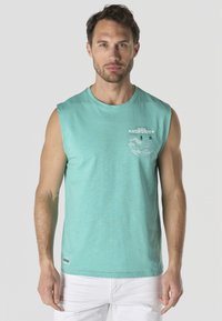 Hombre con camiseta sin mangas turquesa con gráfico blanco y pantalones blancos, de pie contra un fondo gris claro.