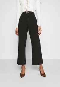 Hochtaillierte schwarze weite Jeans aus Denim mit glatter Textur. Verfügt über einen Knopfverschluss und Seitentaschen. Kombiniert mit spitzen Schuhen im Leopardenmuster.