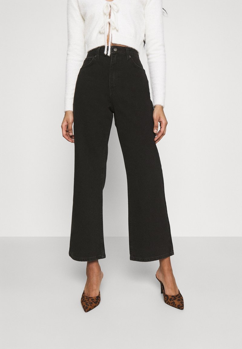Hochtaillierte schwarze weite Jeans aus Denim mit glatter Textur. Verfügt über einen Knopfverschluss und Seitentaschen. Kombiniert mit spitzen Schuhen im Leopardenmuster.