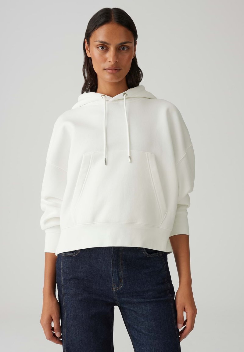 Weißer gekürzter Hoodie mit einer Kängurutasche, Kapuze mit Kordelzug und gerippten Bündchen. Kombiniert mit dunklen Jeans für einen lässigen Look.