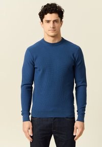 Blauw geribbeld trui met een ronde halslijn en lange mouwen, gemaakt van een gestructureerd breimateriaal. Draagt over donkere denim jeans.