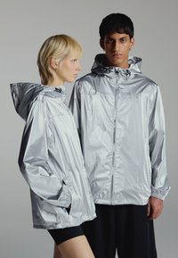 Dos modelos que llevan chaquetas plateadas con capucha y acabado brillante, uno mirando al frente y el otro de perfil con las manos en los bolsillos, sobre un fondo gris liso.