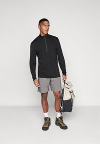 Camisola de manga longa preta com fecho, calções cinzentos, botas de caminhada pretas e castanhas, segurando uma mochila bege e preta, em pé sobre um fundo claro.