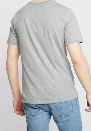 Mężczyzna ubrany w prostą szarą koszulkę z krótkim rękawem i niebieskie dżinsy, pokazany od tyłu na neutralnym tle.