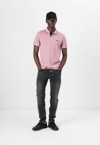 Polo t-shirt i ljusrosa med svarta och vita detaljer, tillsammans med mörkgrå jeans och svarta sneakers. Modellen poserar med händerna i fickorna.