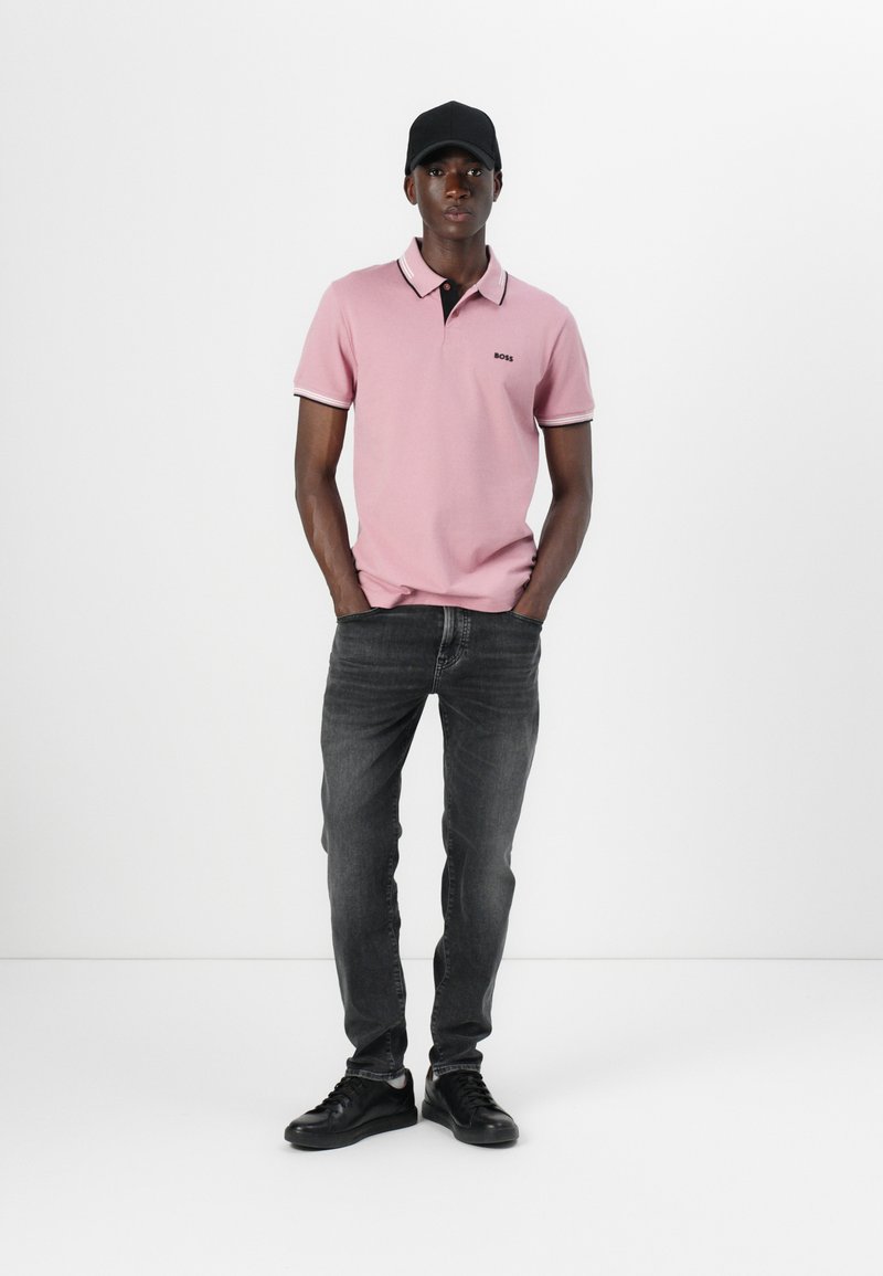 Camisa polo en rosa claro con detalles en negro y blanco, combinada con jeans gris oscuro y zapatillas negras. El modelo posa con las manos en los bolsillos.