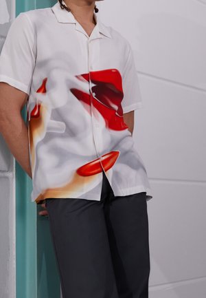 Chemise à manches courtes boutonnée en tissu blanc avec un motif abstrait de lèvres rouges et oranges. Présente une coupe décontractée et un col discret.