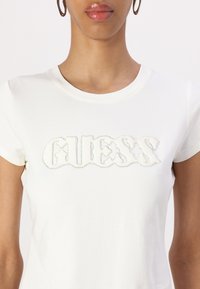Hvit bomulls t-skjorte med en teksturert "GUESS" logo dekorert med rhinsten. Rund hals og kort erm.