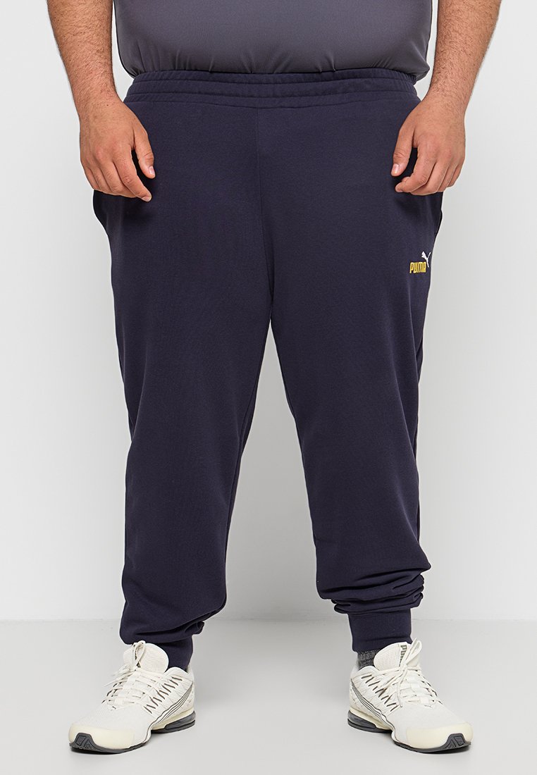 Pantalones de chándal azul marino con cinturilla y puños elásticos, presentan un pequeño logo amarillo de Puma en el muslo izquierdo y una textura suave.