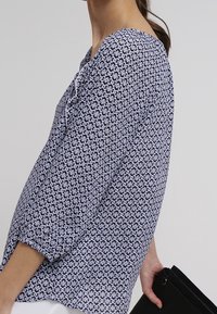 Blouse à motifs bleu marine et blanc avec des manches trois-quarts, en tissu doux et à encolure arrondie, tenue par une personne avec un sac noir.
