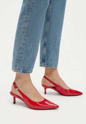 Décolleté in vernice rossa con punta affusolata, dotate di tacco basso e un accento dorato sul cinturino alla caviglia. Indossate con jeans azzurri.
