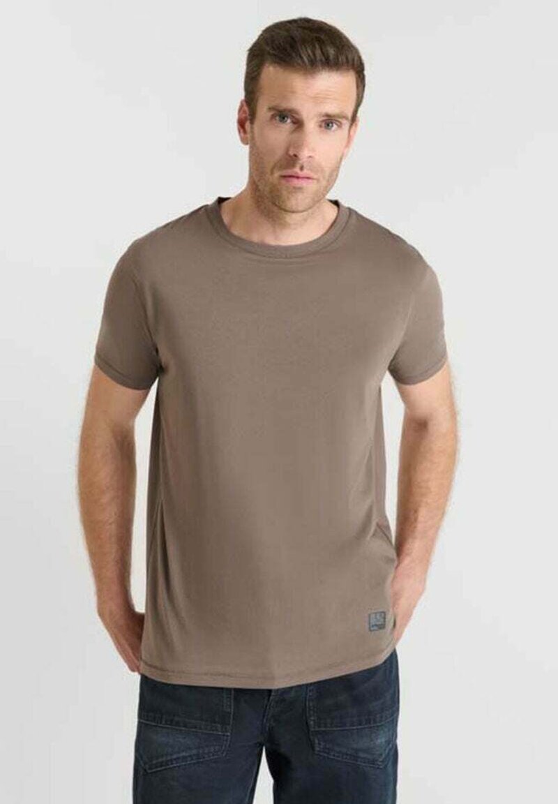 Uomo con capelli corti castani che indossa una semplice maglietta marrone a maniche corte e jeans scuri, in piedi con le mani in tasca contro uno sfondo chiaro.