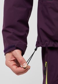 Manica di giacca impermeabile viola con cordino regolabile. Presenta un tessuto texture e un polsino in Velcro. Mano che afferra il toggle del cordino.