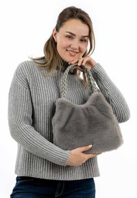 Kobieta w szarym dzianinowym swetrze trzyma dużą szaro-futrzaną torbę z plecionymi uchwytami, uśmiechając się na tle białego tła.