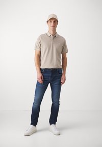 Beige polók, rövid ujjakkal, kék farmernadrág és fehér sportcipő. A sapka a póló színével egyezik, így összehangolt megjelenést teremt.