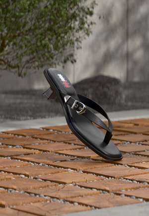 Zwarte sleehak met teenslipper en zilveren gesp, geplaatst op houten tegelvloer buiten met een wazige achtergrond van groen en een muur.