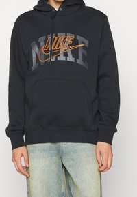 Svart hoodie i bomullsblandning med känguruficka, med stor grå och orange Nike-textlogga på framsidan. Dragsko i huvan ingår.