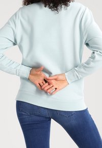 Persoon in lichtblauwe sweatshirt en blauwe jeans, staand met de handen gekruist achter de onderrug, gezien van achteren.