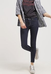 Mujer vestida con jeans skinny oscuros, camiseta negra de Levi's, cárdigan gris abierto y zapatillas rosa claro, de pie con una pierna doblada.