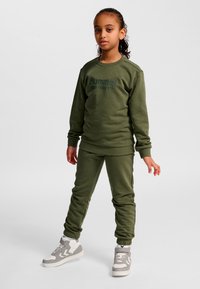 Barn står klädd i olivgrön sweatshirt och byxor med vita och grå sneakers mot enfärgad vit bakgrund.