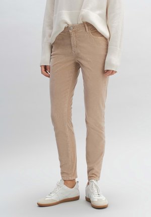 Person trägt beige Slim-Fit-Hosen, weißen Strickpullover und weiße Freizeitsneaker und steht vor einem einfachen hellen Hintergrund.