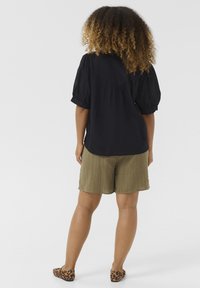 Blusa nera con maniche a sbuffo abbinata a pantaloni corti texture verde oliva e scarpe basse con stampa leopardata, vista da dietro su uno sfondo neutro.