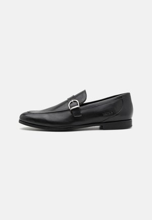 Mocassins en cuir noir avec un bout arrondi, un design sans lanières, une petite boucle décorative et une semelle texturée pour une meilleure adhérence.