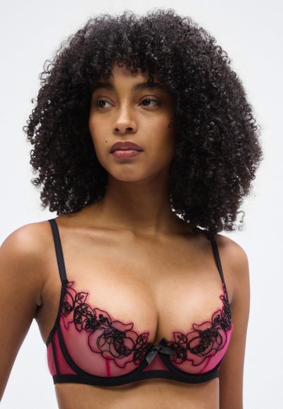 Sostén de encaje rosa translúcido con bordado floral negro, tirantes delgados y detalle de lazo en el centro. Tirantes ajustables y soporte con aro.