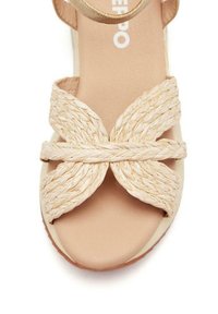 Sandalia beige con diseño de tiras trenzadas formando una X, plantilla acolchada y tirante delgado al tobillo. Textura suave con suela plana.