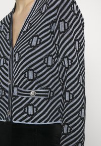 Chaqueta corta con patrón negro y azul, con rayas verticales y motivos geométricos. Presenta un cierre de botón y un bolsillo frontal.