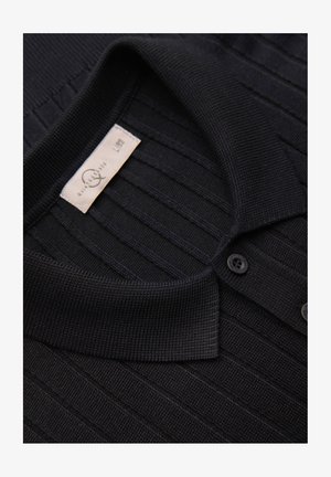 Polo en maille côtelée noire avec col replié, deux boutons et une étiquette visible de taille et d’entretien à l’intérieur de l’encolure.