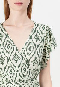 Πράσινο και κρεμ φόρεμα με σχέδιο, με σχεδίου τύπου περιτύλιξης, που διαθέτει κοντά φτεράκια και βαθύ V-decolleté. Ύφασμα με υφή και λουλουδάτο μοτίβο.