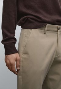 Brown knit sweater with ribbed cuffs above beige trousers featuring side pockets and a flat front design. Smooth texture, classic fit.