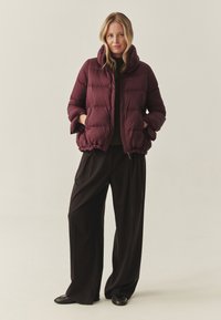 Burgunderfarvet puffjakke med quiltet design, høj krave og elastisk kant. Båret over en mørk sweater og parret med brede sorte bukser.