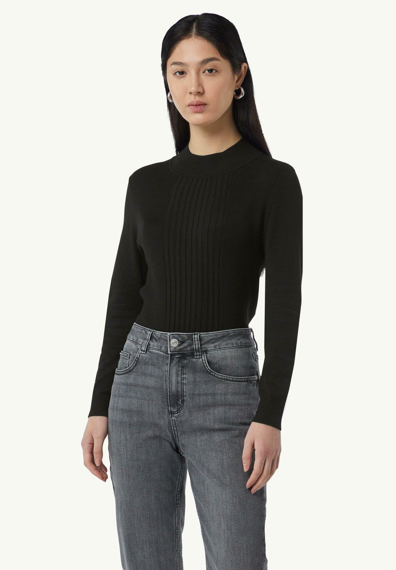 Schwarzer, gerippter Pullover mit hohem Kragen und langen Ärmeln, kombiniert mit grauen Jeans. Der Stoff wirkt glatt und figurbetont.