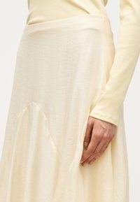 Κοντινό ενός ατόμου που φοράει μια κρεμ ριμπ πλεκτή μπλούζα με μακριά μανίκια και μια κυμαινόμενη ελεφαντόδοντη φούστα με διακριτή υφή, το χέρι ακουμπάει στη φούστα.