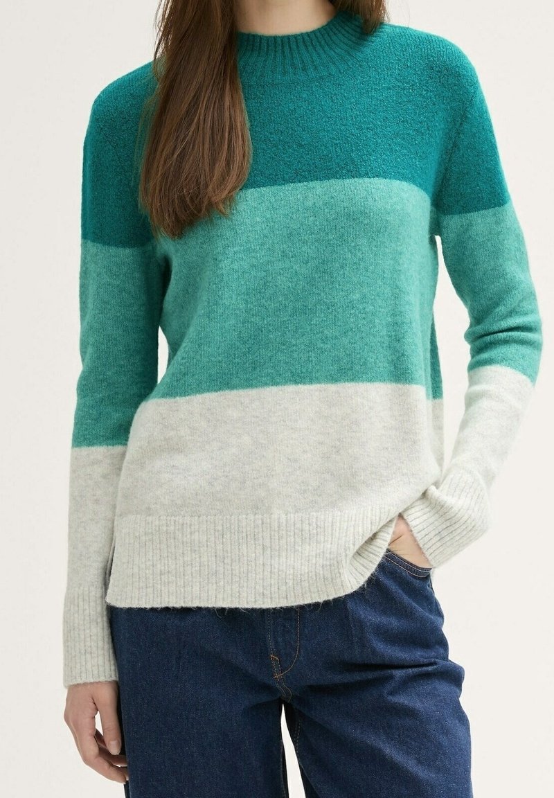 Femme portant un pull en maille manches longues avec des rayures horizontales bleu sarcelle, vert clair et crème et un jean bleu, une main dans la poche.