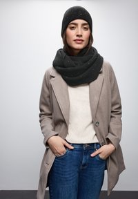 Grå uldfrakke, sort strikket hue, tekstureret sort tørklæde med hvide pletter, cremefarvet sweater og blå jeans, alt sammen med en afslappet pasform.