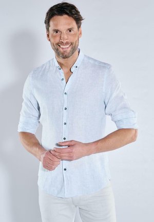 Smiling man with light beard wearing a light blue buttoned shirt with rolled-up sleeves, standing against a plain background.