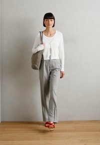 White cropped cardigan over a white top, paired with gray high-waisted trousers. Red strappy sandals, holding a gray tote bag. Light background.