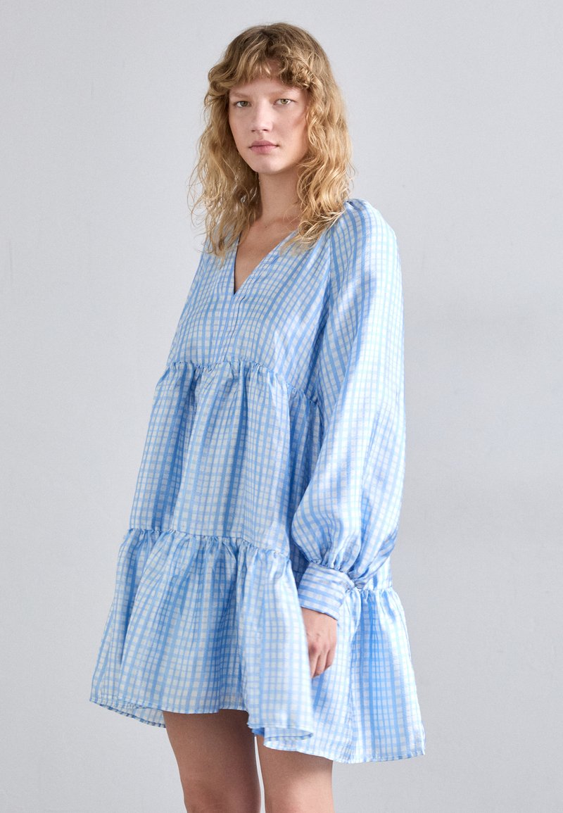 Robe à carreaux bleus et blancs avec décolleté en V, manches longues et ourlet à volants. Fabriquée en tissu léger, elle présente des détails froncés aux poignets.