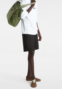 Grüner Stoff-Rucksack mit Trägern, weißes Langarmshirt, schwarze Shorts und braune Wildlederschuhe mit weißen Schnürsenkeln. Einfaches, funktionales Design.