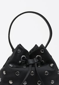 Bolso tipo cubo de tela negra con tachuelas y ojales plateados, cierre con cordón ajustable y asa delgada. Textura suave y diseño minimalista.