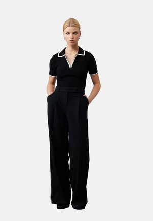 Zwarte polo met witte kraag en rits, gecombineerd met hoge taille zwarte broek. Model staat met handen in de zakken. Gladde texturen.