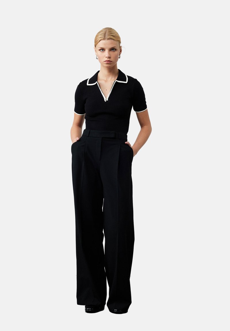 Zwarte polo met witte kraag en rits, gecombineerd met hoge taille zwarte broek. Model staat met handen in de zakken. Gladde texturen.