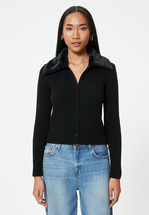 Schwarze gerippte Strickjacke mit einem Kragen aus Kunstfell, mit durchgehender Knopfleiste und langen Ärmeln. Kombiniert mit hellblauen Denim-Jeans.