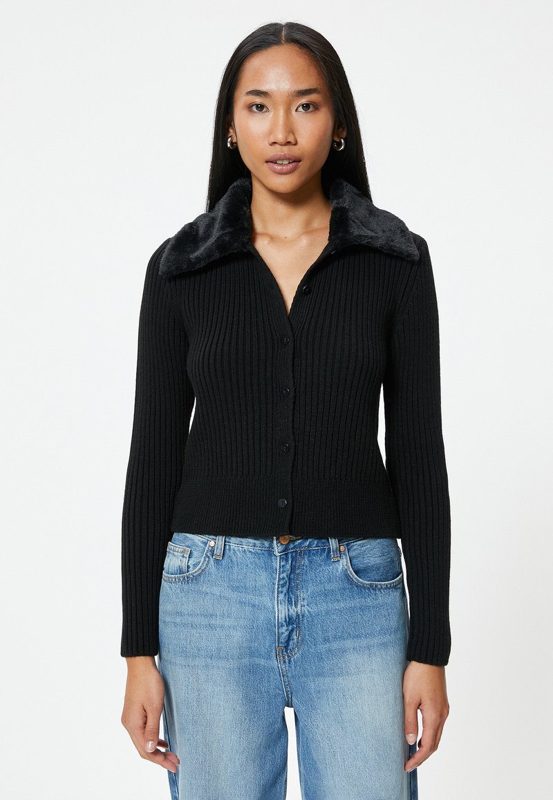 Cardigan nero a coste con colletto in pelliccia sintetica, caratterizzato da una chiusura con bottoni e maniche lunghe. Abbinato a jeans in denim blu chiaro.