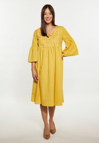 Gelbes Kleid mit langen, ausgestellt geschnittenen Ärmeln und bestickten Details am Ausschnitt. Knielang, aus leichtem Stoff.
