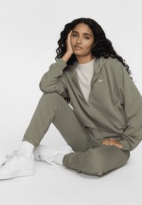 Oliven grønn zip-up hettegenser og matchende joggebukser laget av mykt stoff, paret med en lys grå crew neck t-skjorte og hvite sneakers.