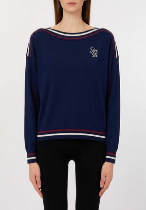 Donna che indossa un maglione blu navy con rifiniture a righe rosse e bianche sul collo, sui polsini, sull'orlo e un piccolo logo ricamato sul petto.